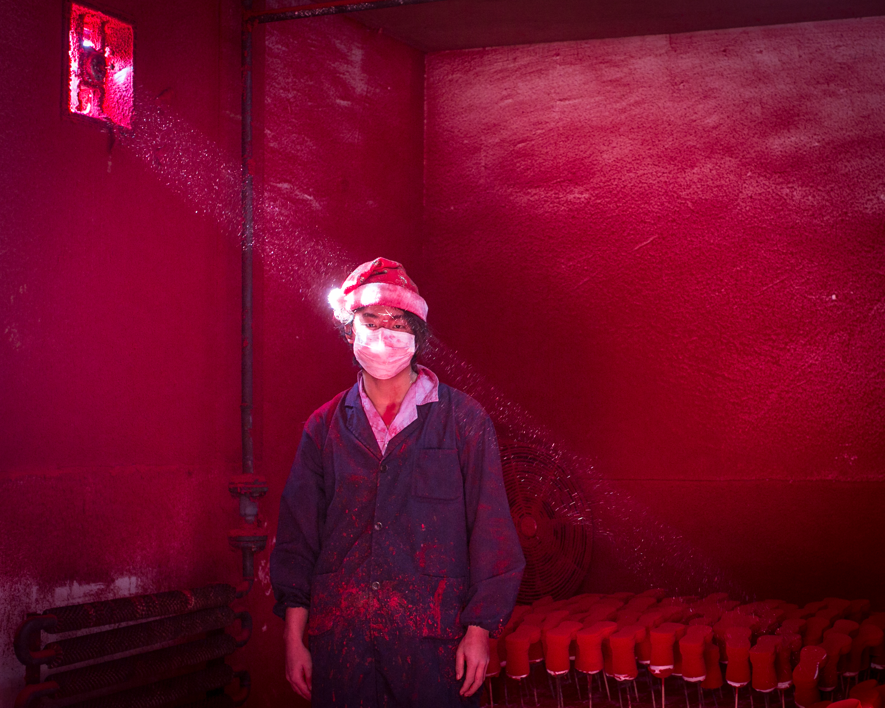 Ronghui Chen, Chine, City Express. Wei, travailleur chinois de 19 ans, porte un masque et un chapeau de père Noël, debout à côté des décorations de Noël qui sèchent dans une usine où la poudre rouge utilisée comme colorant demeure en suspension dans l’air. Il porte six masques par jour, et le chapeau protège ses cheveux de la poussière rouge qui, après plusieurs heures de travail, couvre les travailleurs de la tête aux pieds comme s’il s’agissait de la suie.